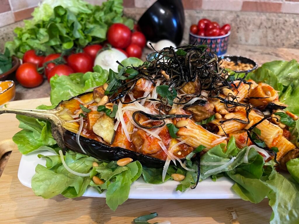 an eggplant boat stuffed with rigatoni alla Siciliana, place in a white dish decorated with lettuce. Behind the dish a display of tomatoes, eggplants, and lettuce.