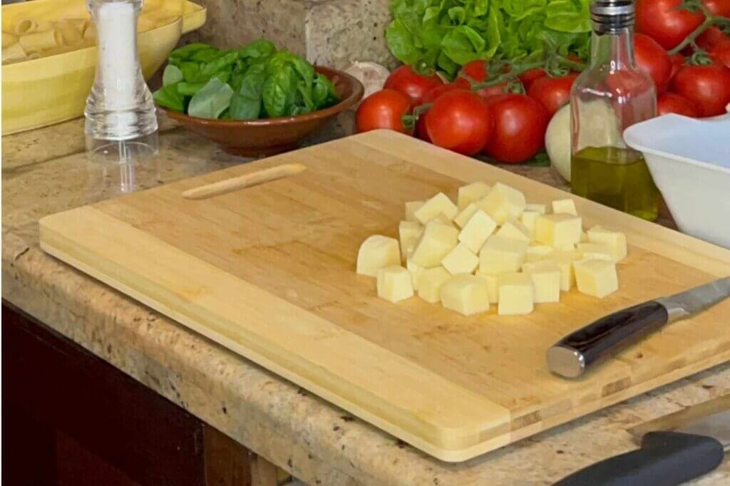 Cubed low-moisture mozzarella cheese prepared for Rigatoni Alla Siciliana on a wooden cutting board with fresh Italian ingredients