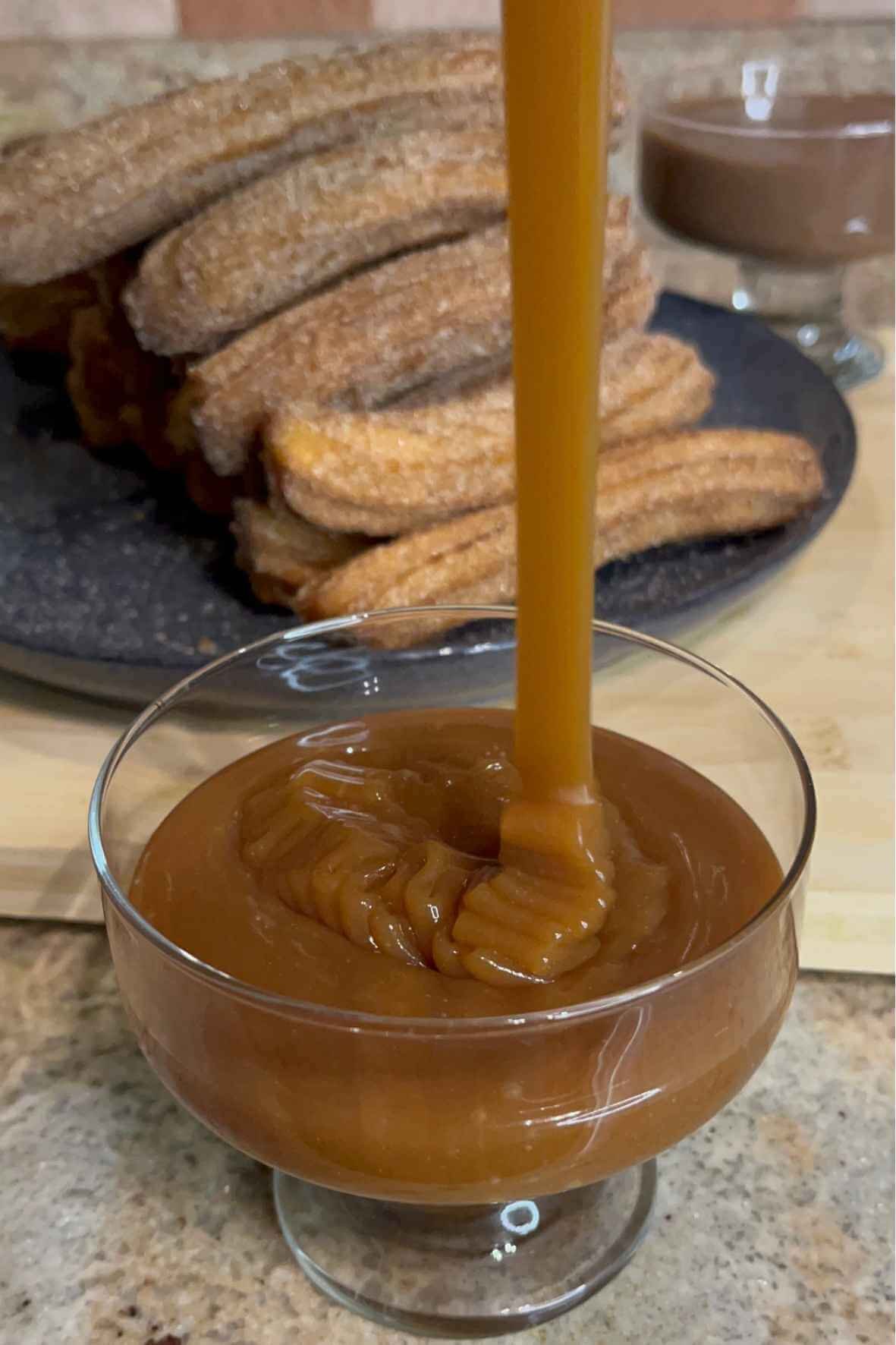 Homemade caramel sauce being poured into a glass dessert bowl with cinnamon sugar churros in the background