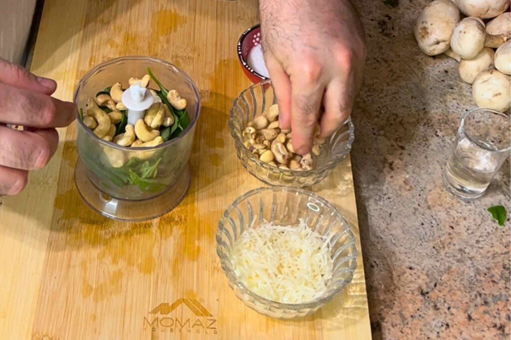 Adding cashews to a food processor with fresh basil and grated Parmesan to prepare homemade pesto sauce.