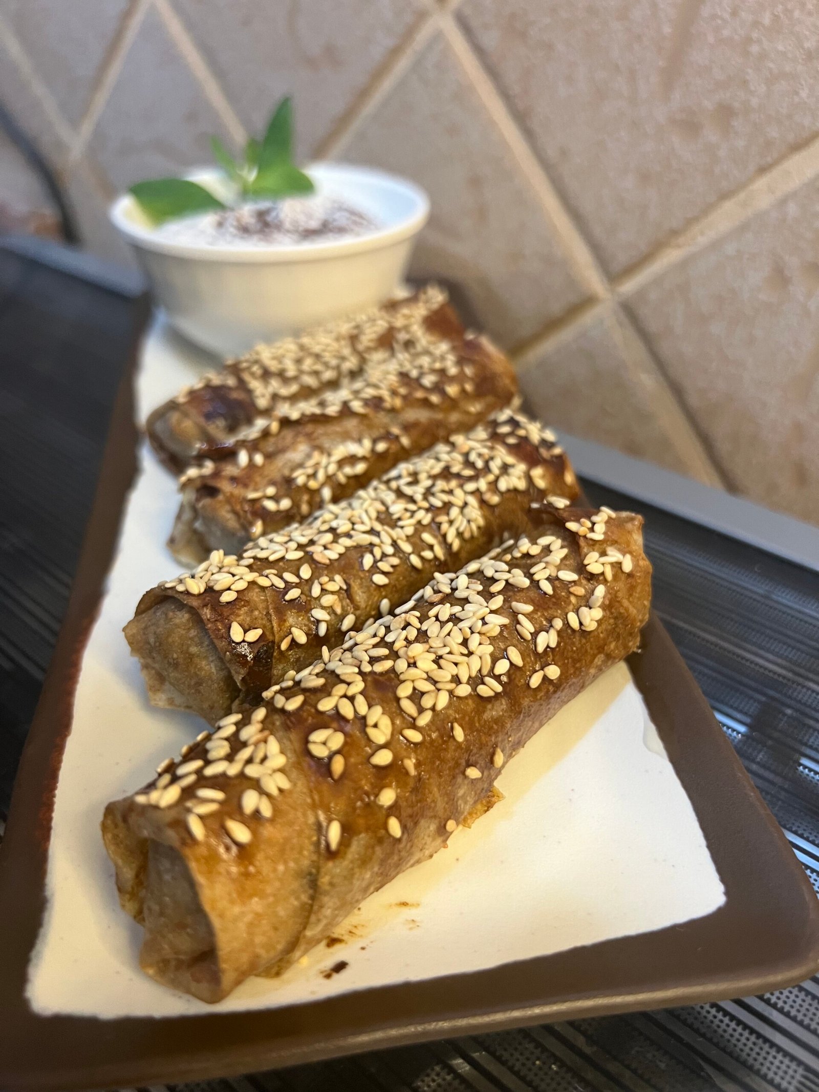Four musakhan rolls sprinkled with sesame seeds, served on a white plate with a small bowl of yogurt on the side.