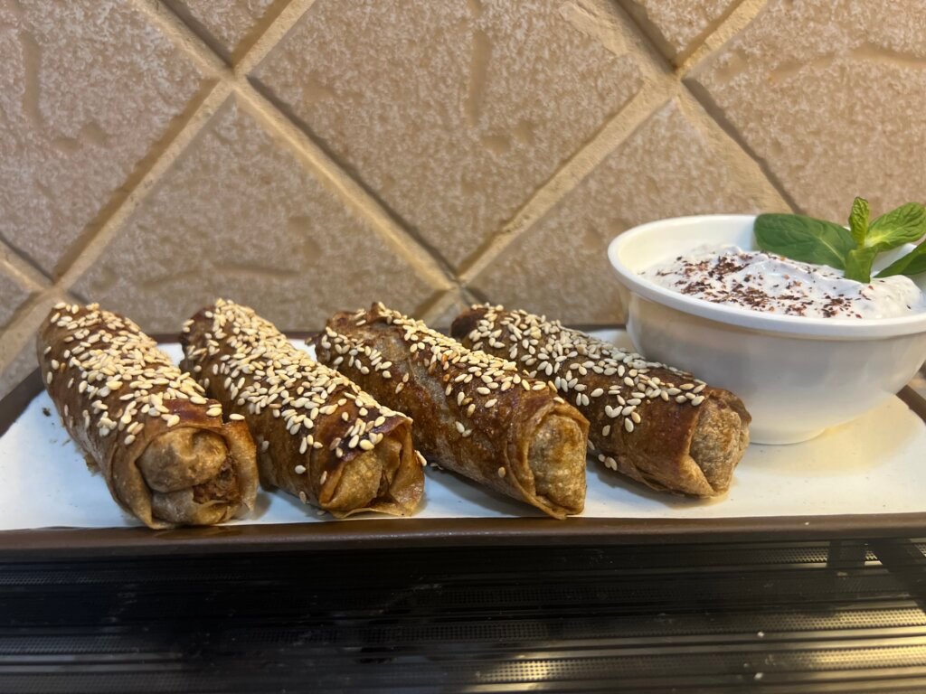 Four musakhan rolls sprinkled with sesame seeds, served on a white plate with a small bowl of yogurt on the side.