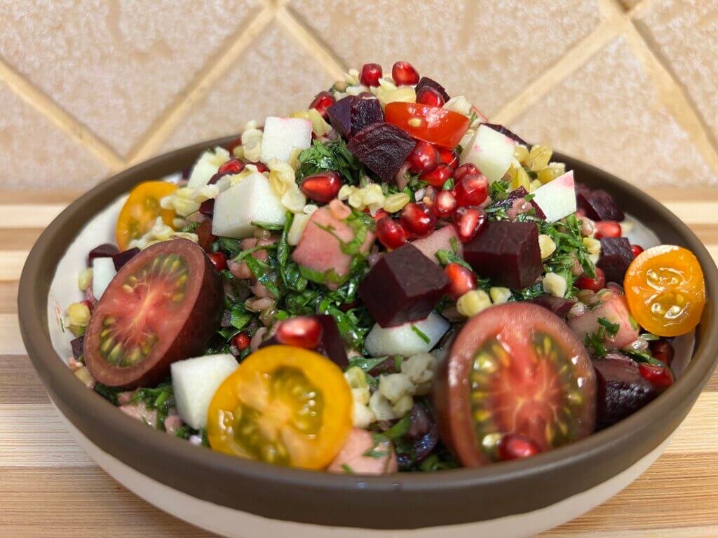 Close-up of a colorful freekeh tabbouleh salad with parsley, pomegranate seeds, beetroot, cherry tomatoes, and diced green apple served in a bowl.