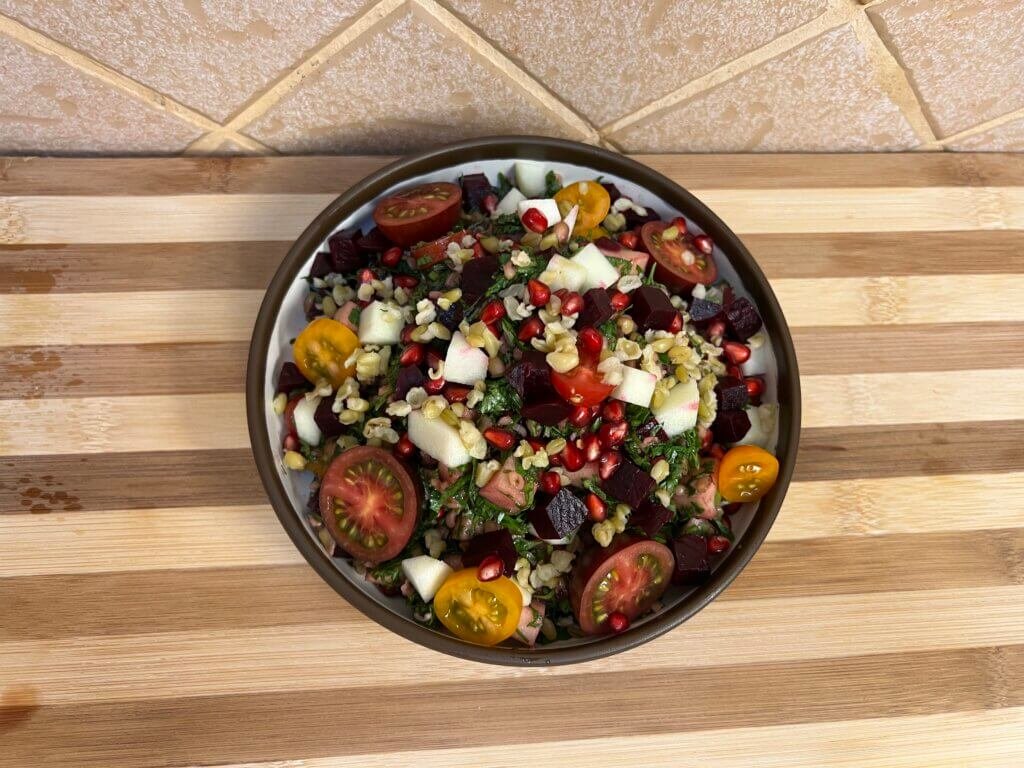 Close-up of a colorful freekeh tabbouleh salad with parsley, pomegranate seeds, beetroot, cherry tomatoes, and diced green apple served in a bowl.
