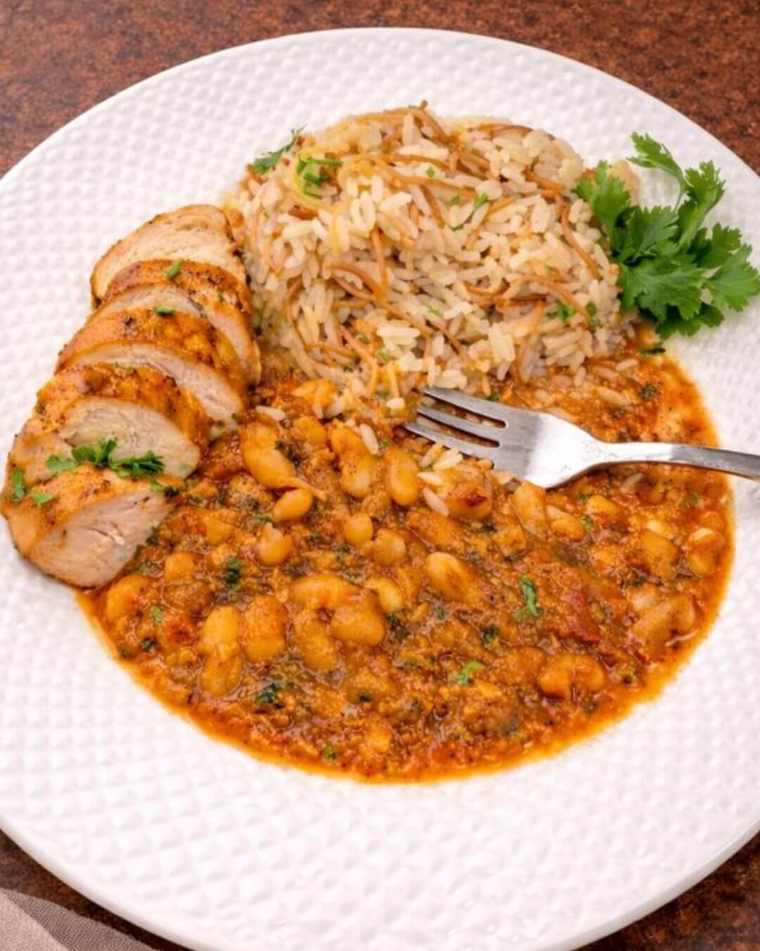 Plate of sliced chicken breast served with white beans stew in tomato sauce and vermicelli rice, garnished with fresh herbs.