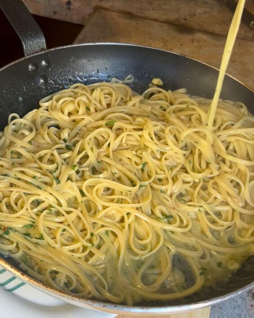 Aglio e olio linguine cooking in a skillet with garlic, olive oil, parsley, and pasta water forming a creamy sauce