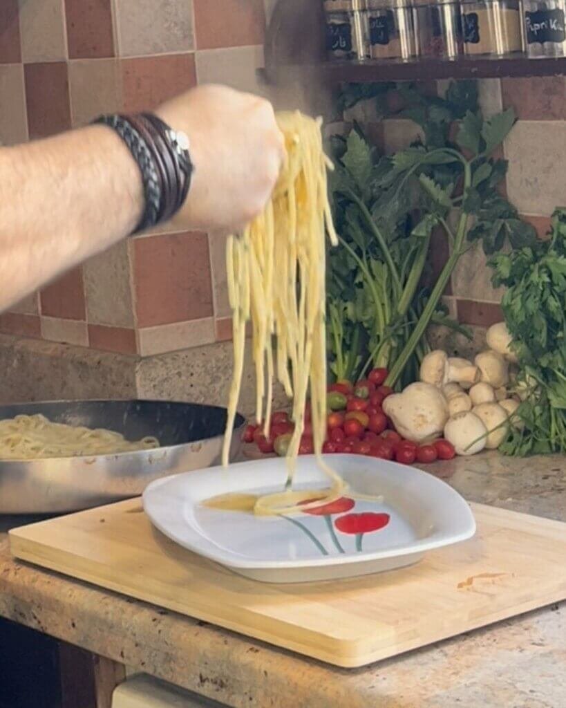 Chef plating aglio e olio linguine pasta onto a white dish in a home kitchen with fresh herbs and vegetables in the background