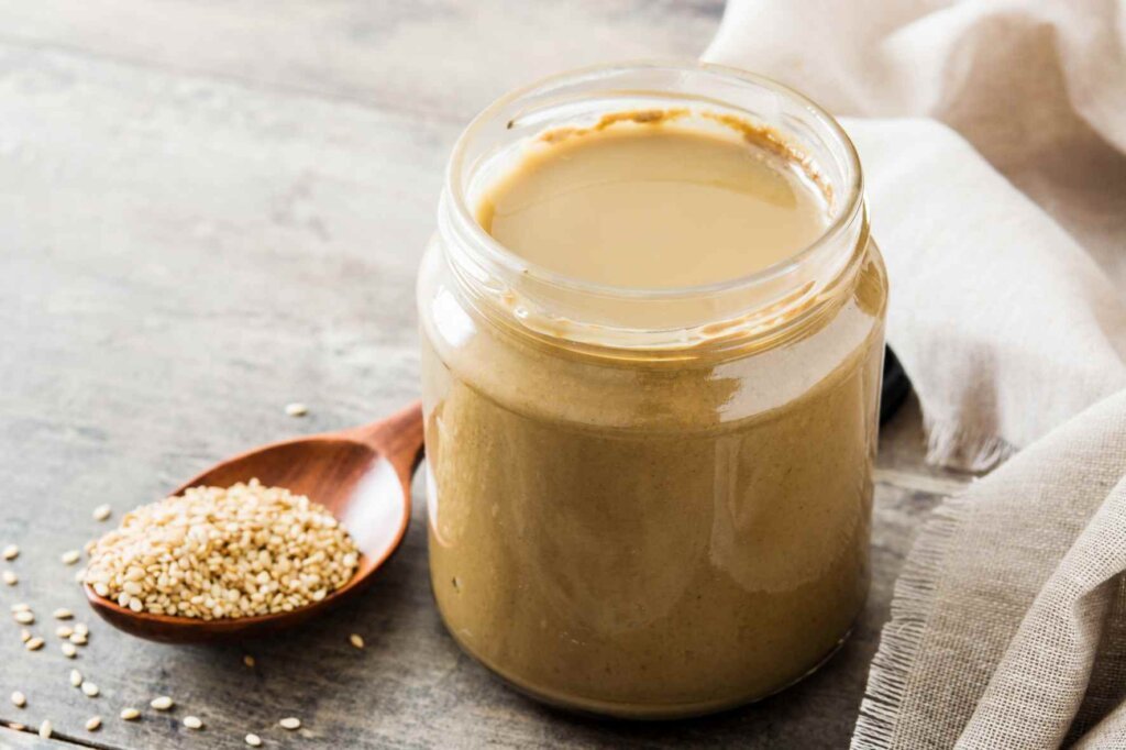 Open jar of creamy tahini beside a wooden spoon filled with sesame seeds.