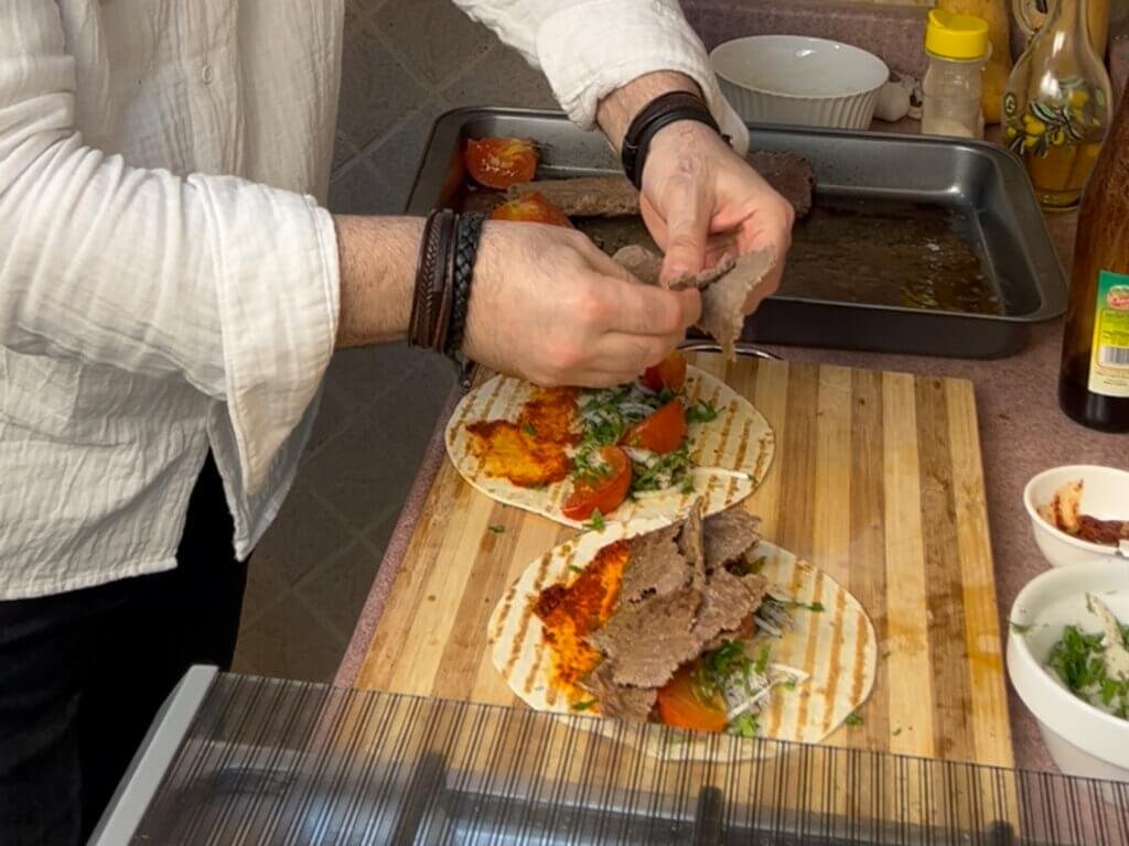 Hands assembling homemade doner kebab wraps with sliced beef, vegetables, and sauce on tortilla bread