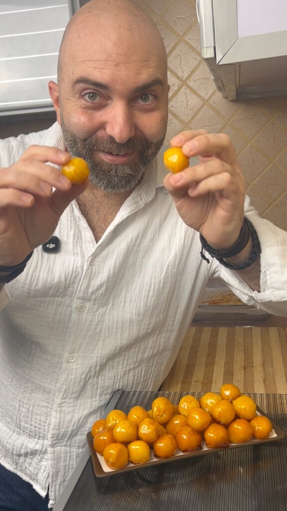 chef holding two bala baiana coconut candies with a tray of caramel coated sweets in the background