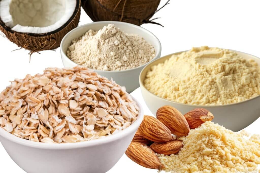Bowls of gluten-free ingredients including oats, almond flour, coconut flour, and corn flour arranged together on a white background.