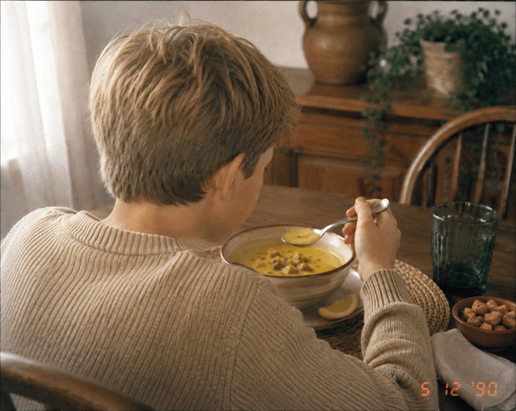 Vintage-style photograph of young François as a boy, seen from behind while eating a bowl of yellow lentil soup topped with croutons, with a lemon wedge on the side.