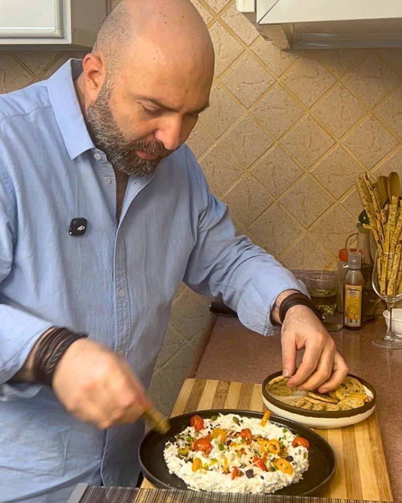 Chef dipping a cracker into creamy feta dip topped with tomatoes, seeds, and herbs in a home kitchen