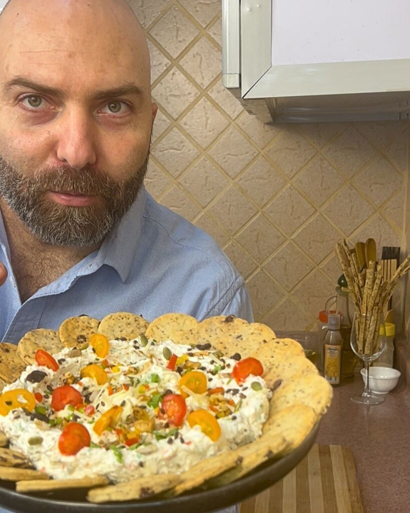 Chef holding a bowl of creamy feta dip topped with cherry tomatoes, seeds, and herbs, served with crackers