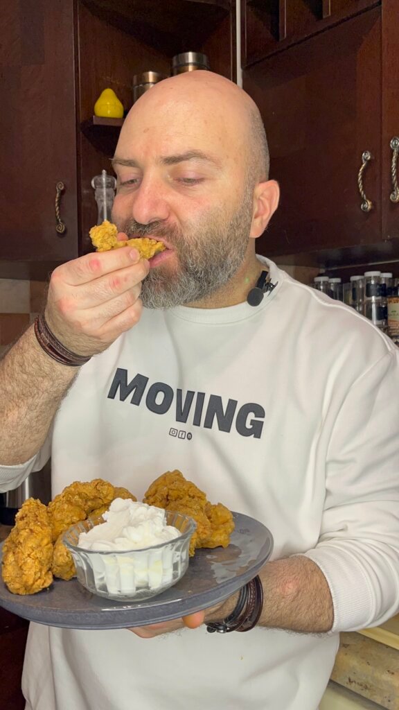 Man eating crispy fried chicken while holding a plate with garlic dip in a kitchen setting