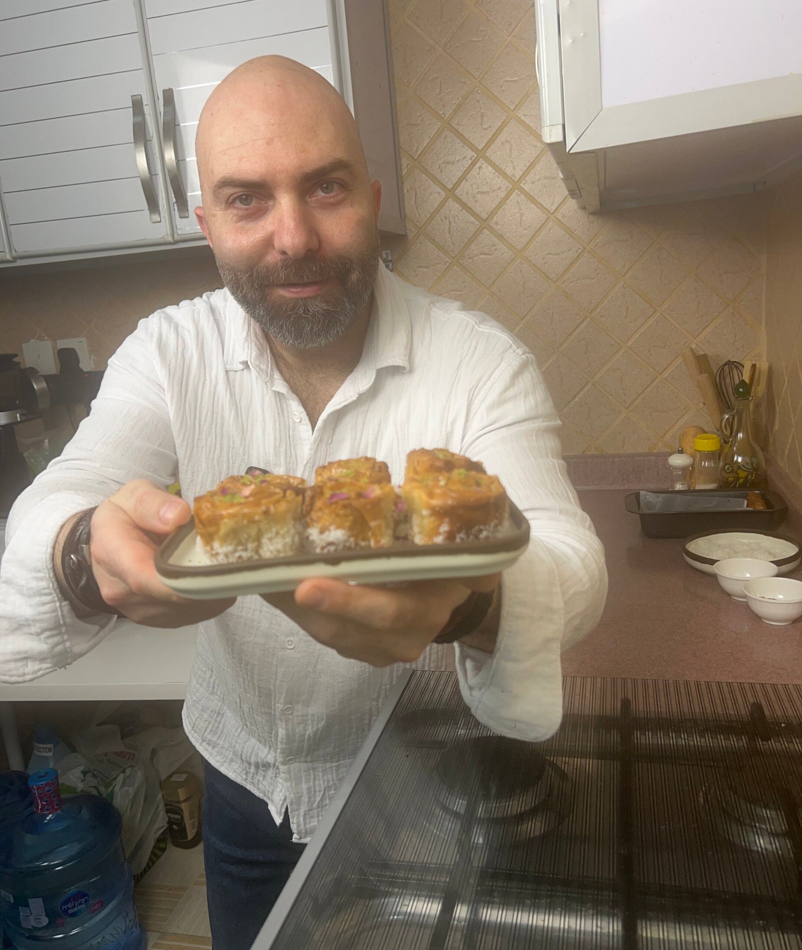 Man holding a plate of baklava roses in a kitchen, showing golden pastries filled with almonds and coconut
