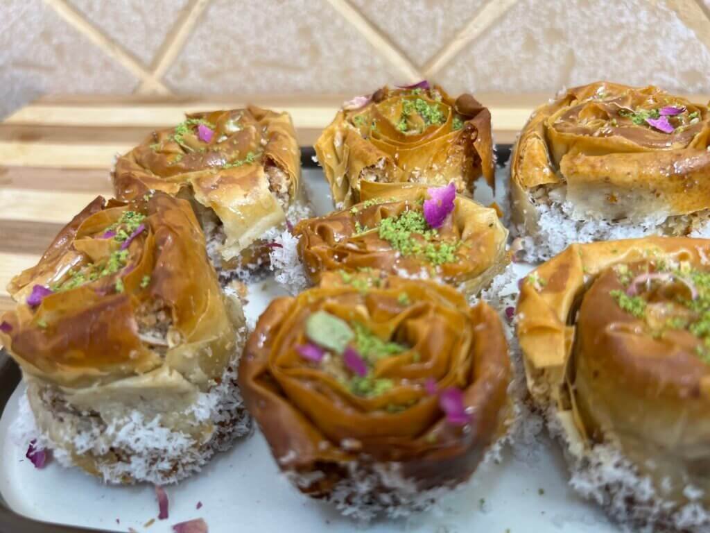 Close-up of golden baklava roses filled with almonds and coconut, topped with crushed pistachios and rose petals on a white plate