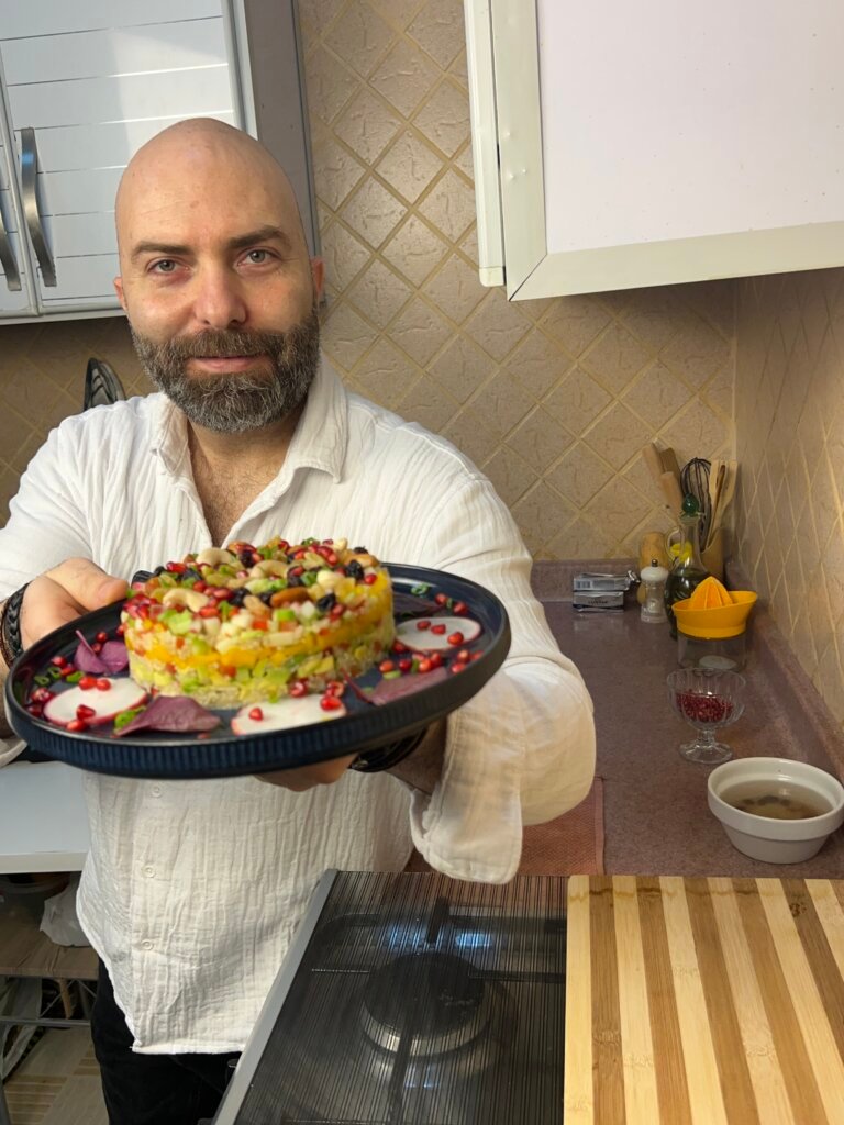 Chef François holding a plate of healthy quinoa salad layered with fresh vegetables, fruits, nuts, and pomegranate seeds in a kitchen
