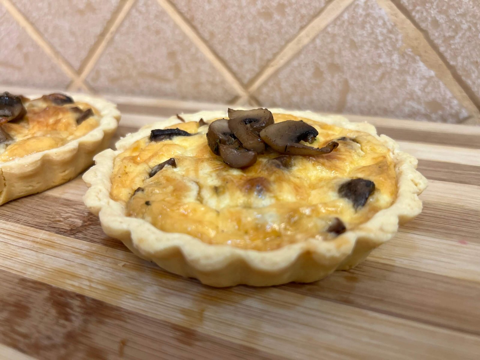 Close-up of a mini mushroom quiche with a golden crust, creamy filling, and sautéed mushrooms on top, placed on a wooden board.