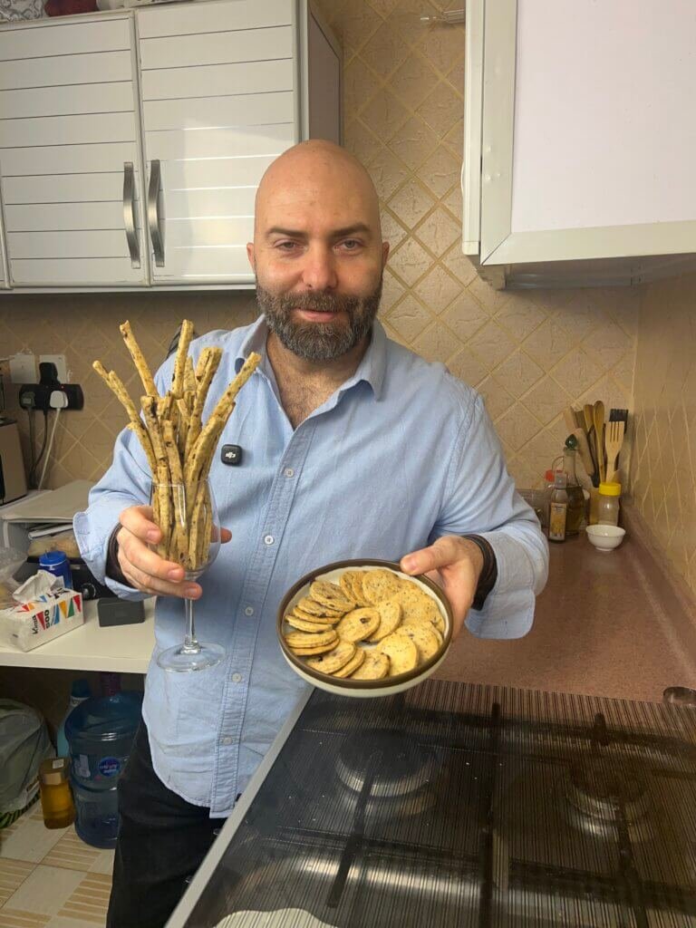 Chef holding thyme and olives breadsticks in a glass in one hand and crispy crackers on a plate in the other