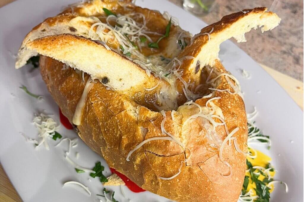 French onion soup served inside a crusty artisan bread bowl, topped with melted cheese and grated Gruyère, presented on a white plate with fresh parsley garnish.