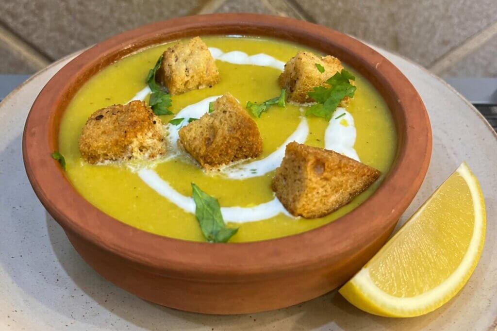 Creamy yellow lentil soup served in a rustic clay bowl on a ceramic plate, topped with crunchy homemade croutons, a swirl of cream, fresh herbs, and a wedge of lemon on the side.