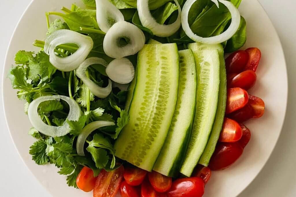 A colorful plate of fresh vegetables featuring cucumber slices, cherry tomatoes, onion rings, leafy greens, and fresh herbs served as a side dish.