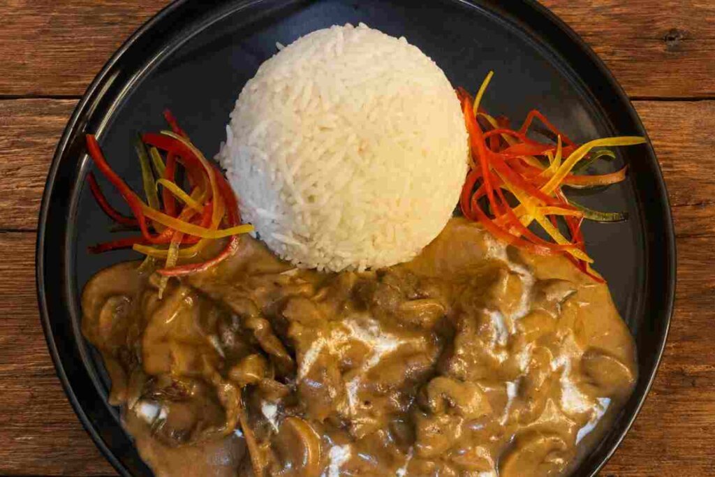 Beef stroganoff recipe with creamy mushroom sauce, white rice, and colorful vegetables on a wooden kitchen table