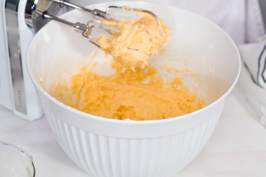 Electric hand mixer beating butter and sugar in a large white mixing bowl to create a light and creamy mixture for cake batter.