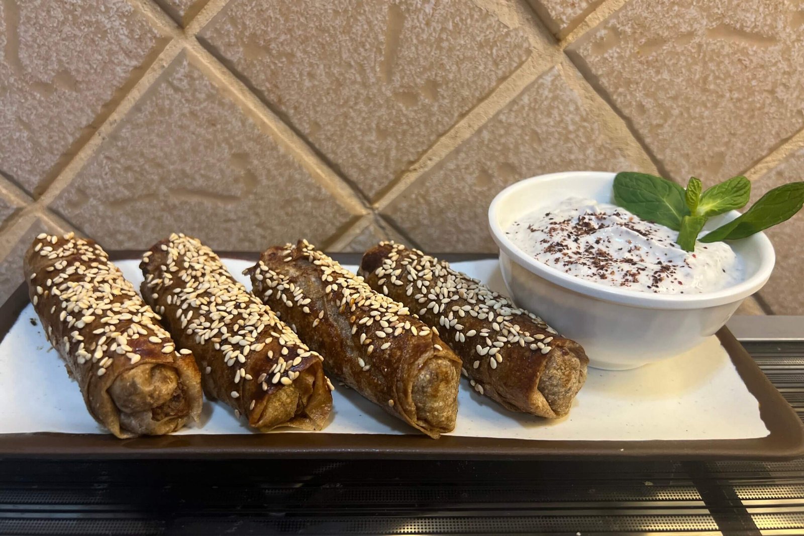 Four musakhan rolls sprinkled with sesame seeds, served on a white plate with a small bowl of yogurt on the side.