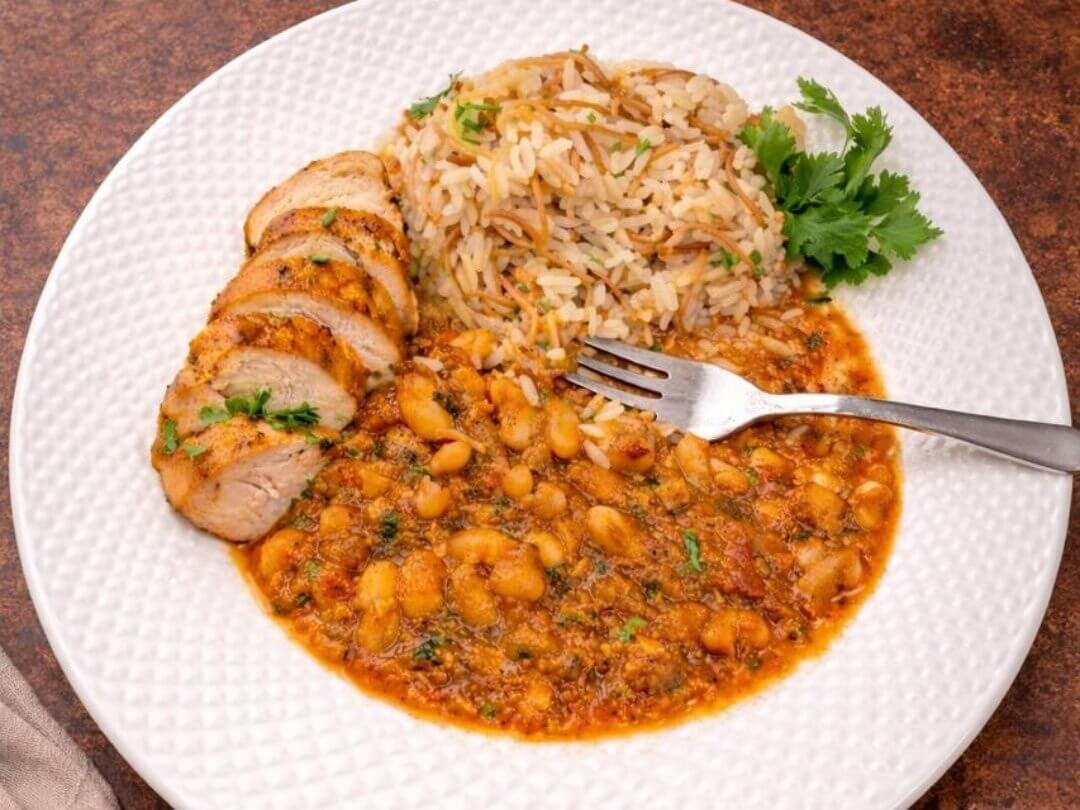 Plate of chicken breast slices served with white beans stew in tomato sauce and vermicelli rice, garnished with fresh coriander.
