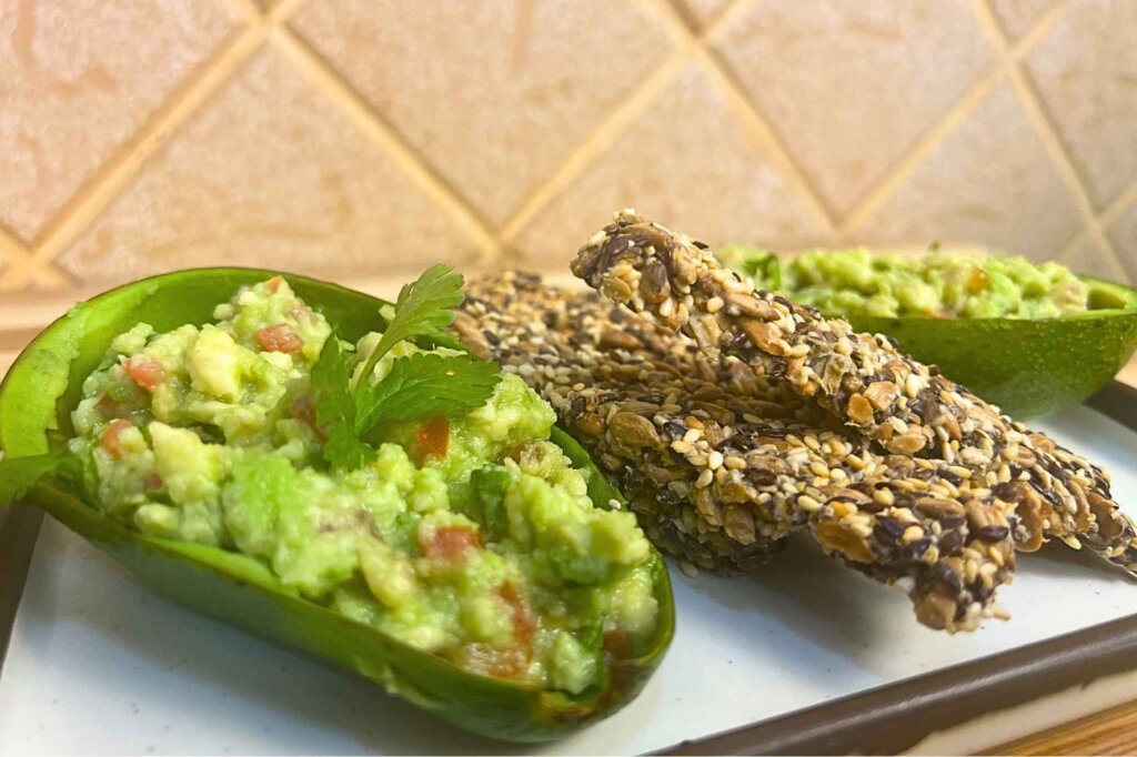 Close-up of homemade gluten-free seed crackers served with fresh avocado guacamole in avocado shells on a white plate.