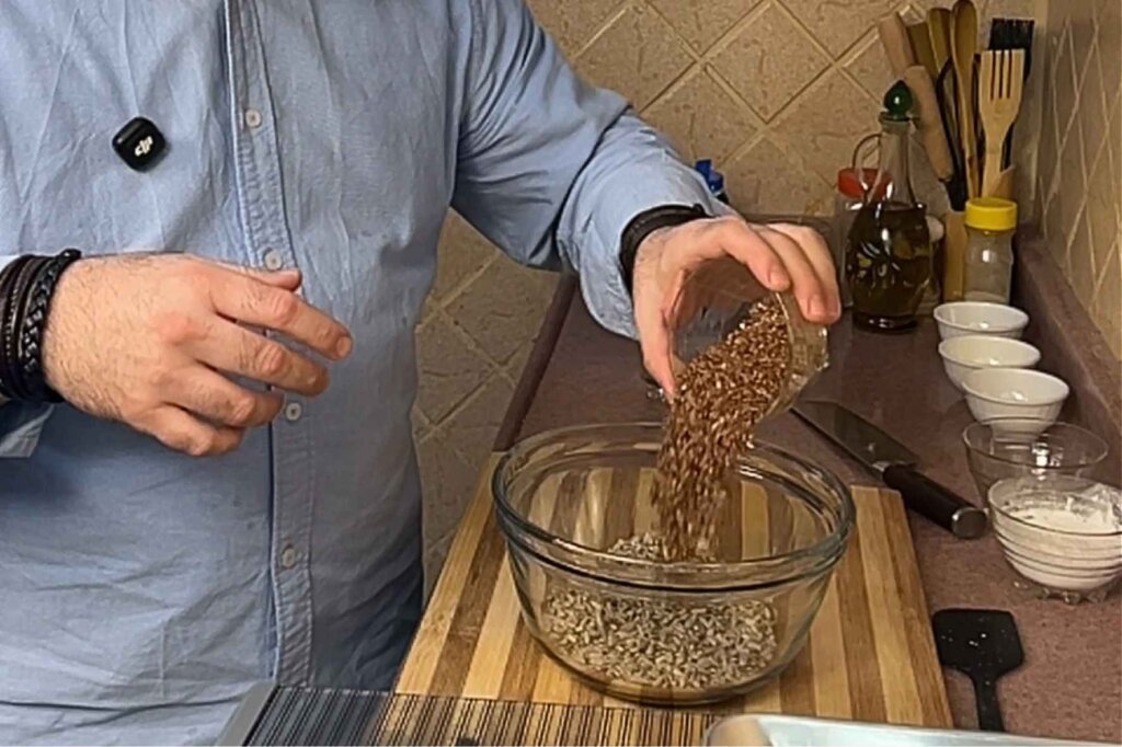 A chef pours flaxseeds into a glass bowl filled with mixed seeds while preparing homemade gluten-free seed crackers in a kitchen.