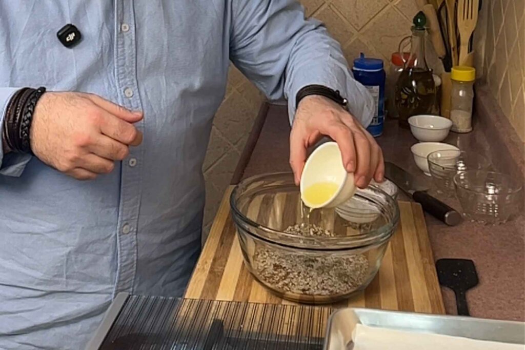 A chef pours oil into a bowl of mixed seeds while preparing homemade gluten-free seed crackers in a kitchen.