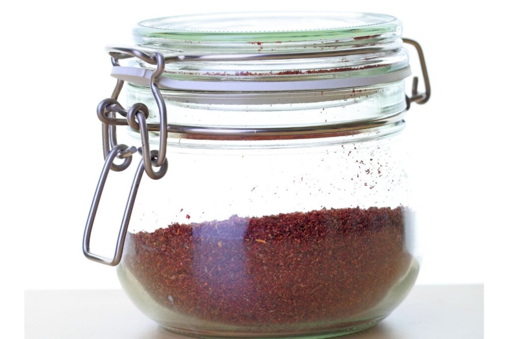 Glass jar filled with freshly ground dark red sumac spice on a white background.