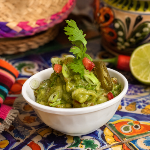 Bowl of fresh guacamole garnished with cilantro, jalapeño, tomatoes, and green onions, displayed on a colorful Mexican-style table setting.