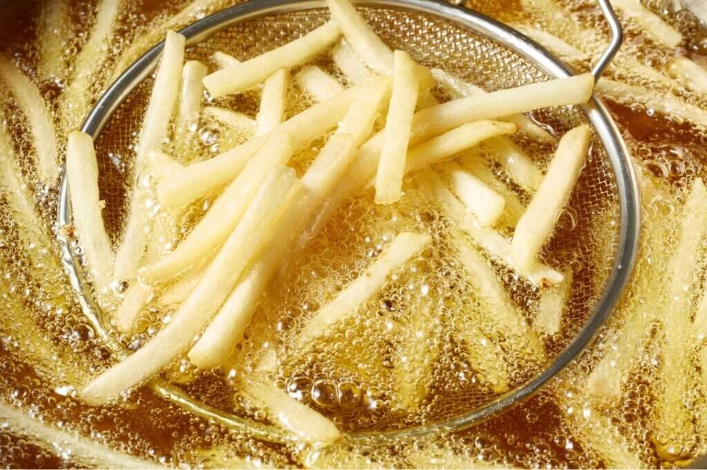 Fresh-cut French fries frying in hot oil inside a deep fryer, surrounded by bubbling oil, with a wire skimmer lifting a batch of potatoes.