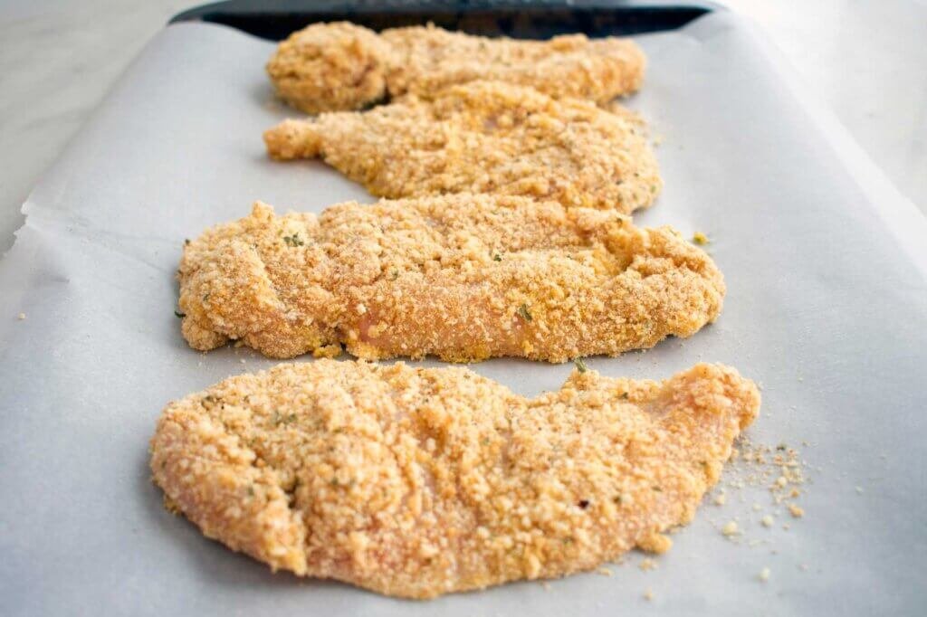 Breaded chicken cutlets and drumsticks resting on a parchment-lined tray before frying, allowing the coating to adhere firmly for a crisp, even crust.