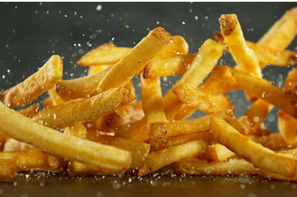 Golden French fries being sprinkled with salt immediately after frying, the ideal time to season them while preserving a crispy texture.