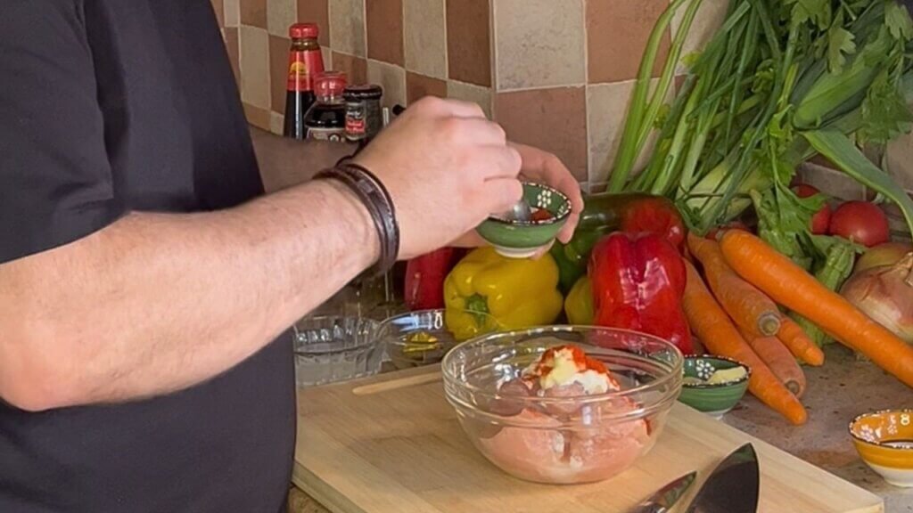 Chef seasoning raw chicken in a bowl with yogurt and spices while preparing a marinade surrounded by fresh vegetables in a kitchen.