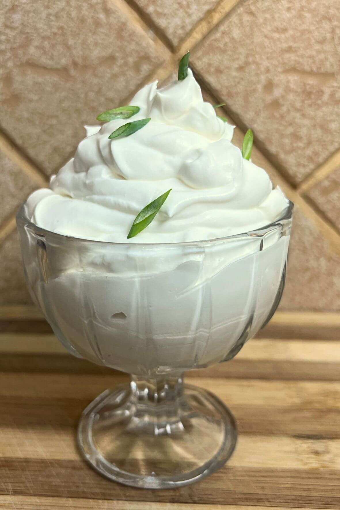 Close-up of creamy homemade sour cream swirled into soft peaks in a glass dessert bowl, garnished with sliced green onions and served on a wooden board against a warm kitchen tile background.