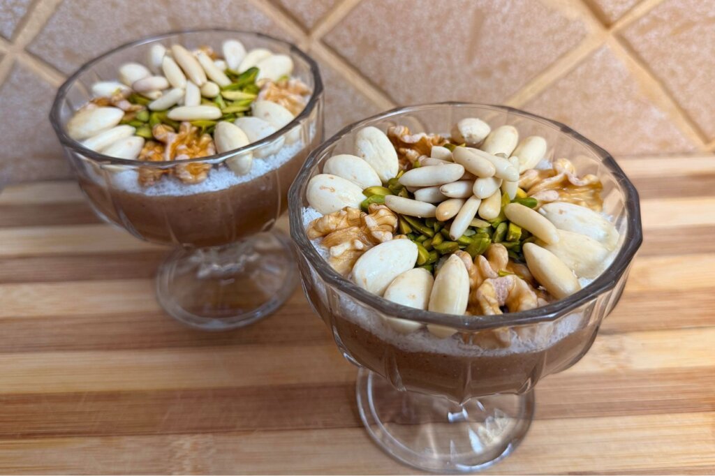 Two glass dessert cups filled with Lebanese meghleh and topped with shredded coconut, almonds, pistachios, walnuts, and pine nuts.