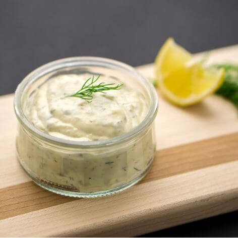 Creamy homemade tartar sauce in a glass jar garnished with fresh dill, served with lemon wedges on a wooden board.