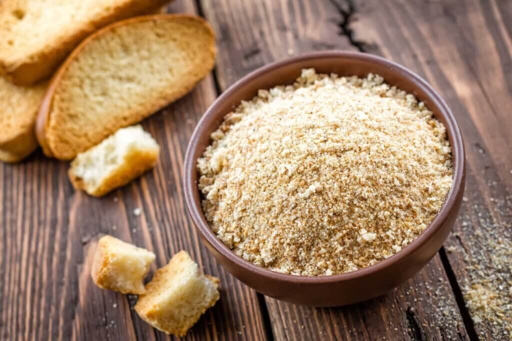 Bowl filled with finely ground traditional breadcrumbs surrounded by slices of toasted bread and bread cubes on a rustic wooden table.
