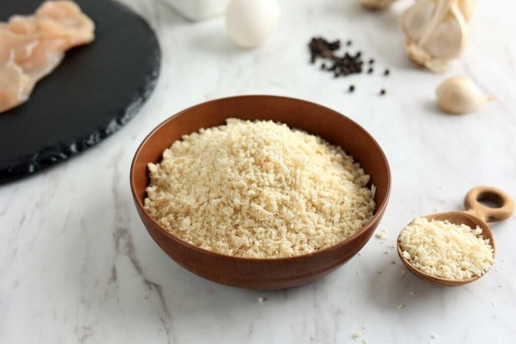 Bowl filled with Japanese panko breadcrumbs showing their large, flaky texture, with raw chicken, garlic, and seasonings in the background.