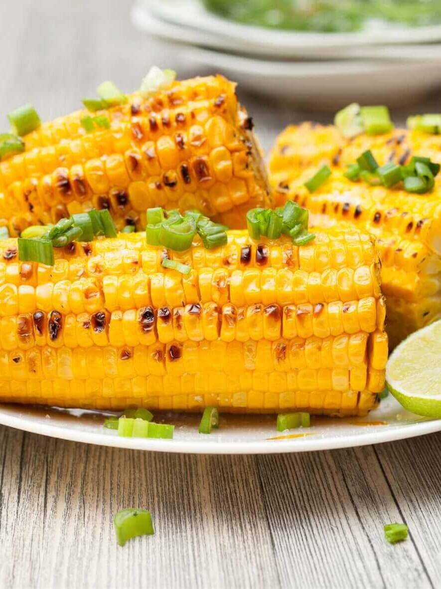 Grilled corn on the cob served with lime wedges and chopped green onions on a white plate, a delicious side dish for fish sticks and other seafood meals.