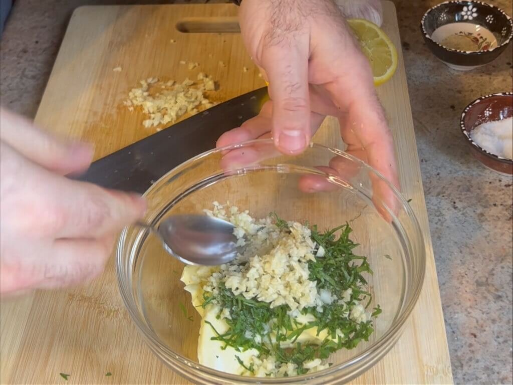 Preparing homemade garlic butter with fresh garlic, parsley, butter, and lemon juice in a glass bowl on a wooden cutting board.