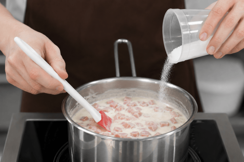 Chef adding salt to a creamy ham sauce while stirring in a saucepan during a cooking lesson about proper seasoning.