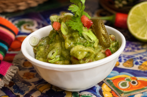 Bowl of fresh guacamole garnished with cilantro, jalapeño, tomatoes, and green onions, displayed on a colorful Mexican-style table setting.