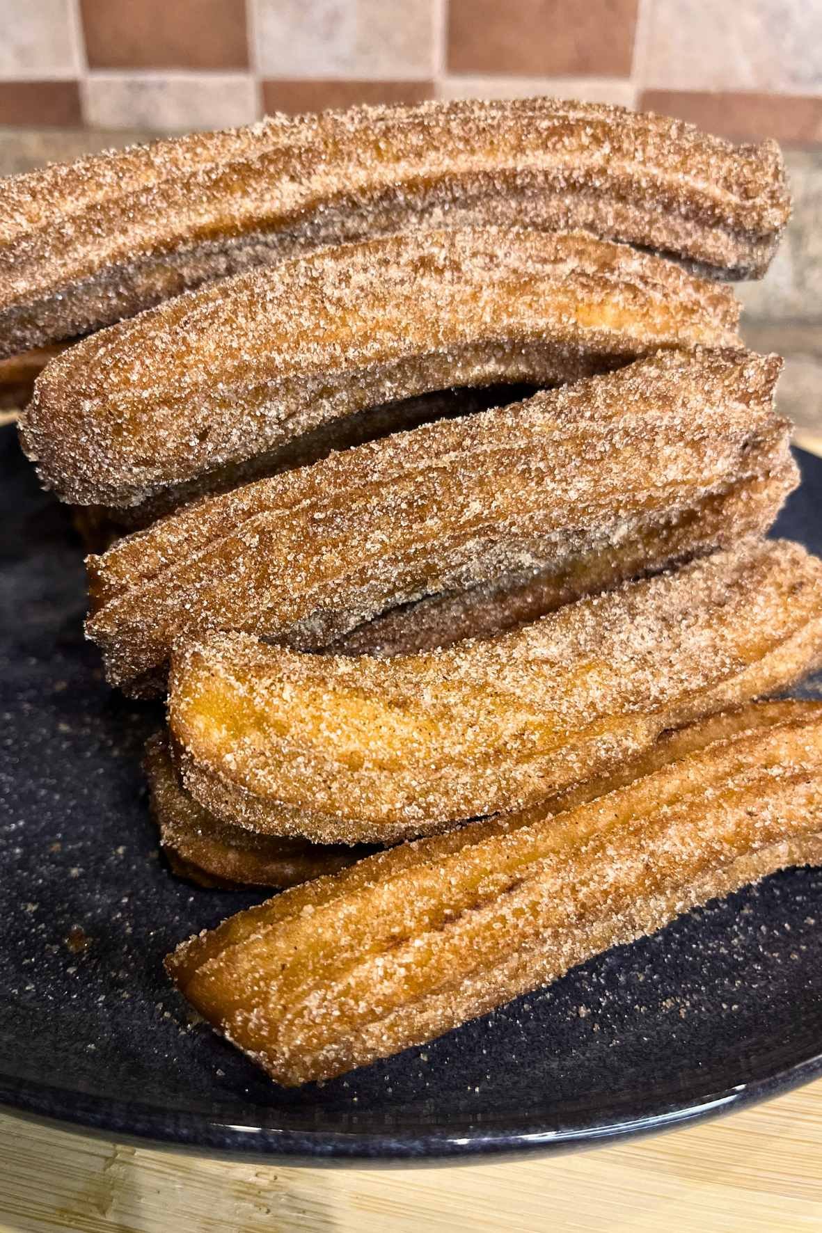 Golden homemade churros coated in cinnamon sugar, stacked on a black plate and served warm.